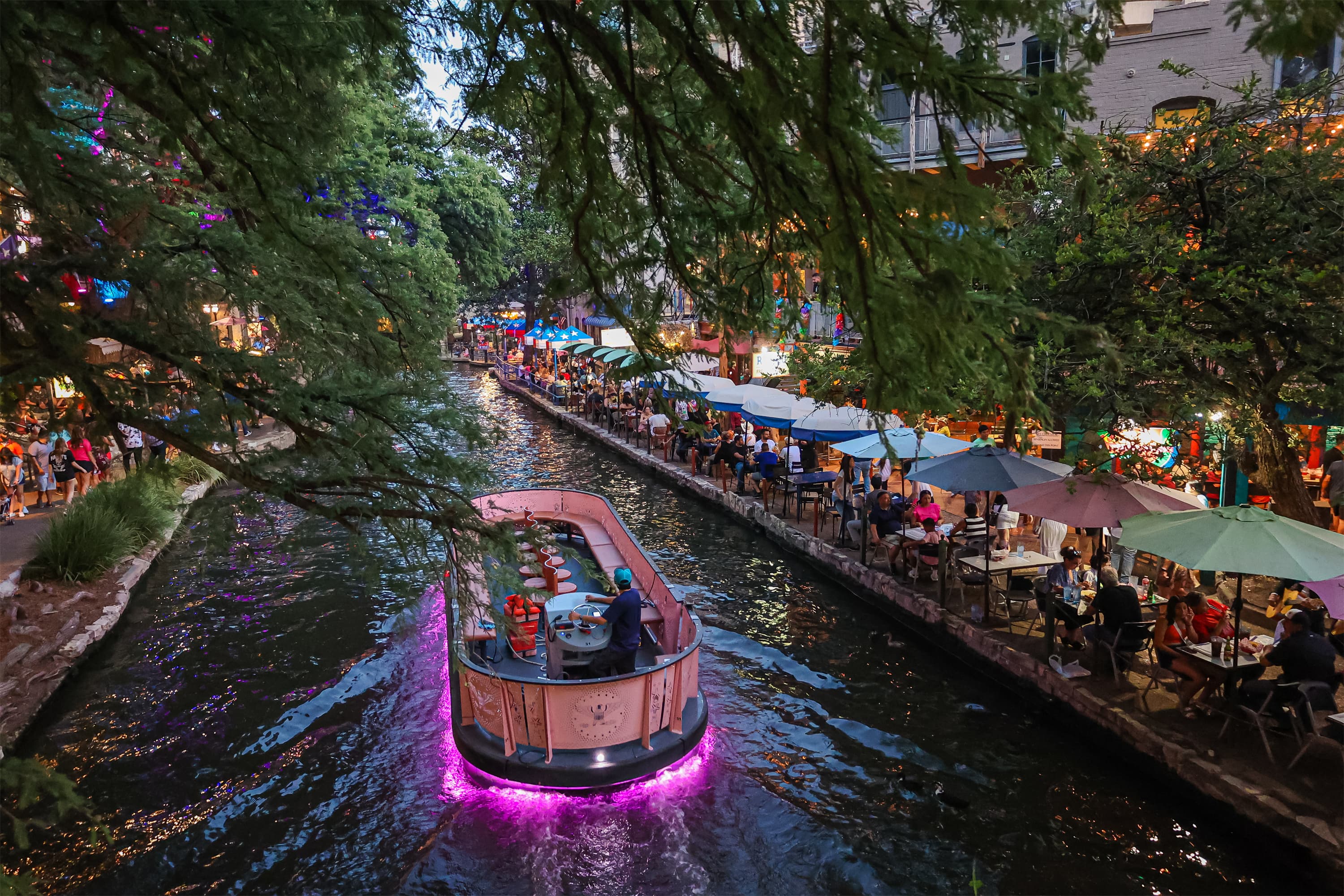 A lit barge moving along the San Antonio River Walk past riverside diners at dusk